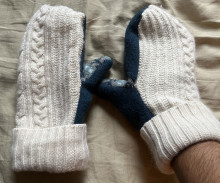 A picture of handmade mittens on a tan fabric. The mittens are white and blue with a combination of knit fabric and fleece.